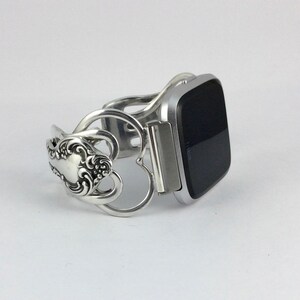 Puede incluir: Pulsera de cuchara de plata con un dise&ntilde;o en forma de coraz&oacute;n, que sostiene un reloj inteligente plateado con una pantalla negra.