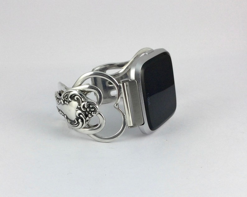 Puede incluir: Pulsera de cuchara de plata con un dise&ntilde;o en forma de coraz&oacute;n, que sostiene un reloj inteligente plateado con una pantalla negra.