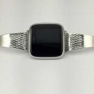 K&ouml;nnte beinhalten: Ein silbernes Metallarmband mit einem Wabenmuster. Das Armband ist an einer silbernen Fitbit-Smartwatch mit schwarzem Bildschirm befestigt.