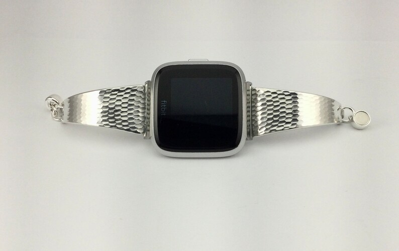 K&ouml;nnte beinhalten: Ein silbernes Metallarmband mit einem Wabenmuster. Das Armband ist an einer silbernen Fitbit-Smartwatch mit schwarzem Bildschirm befestigt.