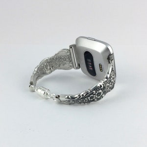 May include: Silver Fitbit watch with a decorative floral spoon bracelet. The bracelet is made from repurposed silverware and features intricate floral patterns. The watch is silver and has a black screen. The bracelet is connected with a magnetic clasp.