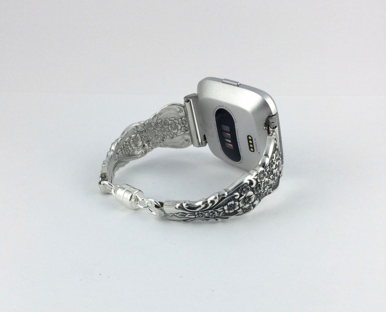 May include: Silver Fitbit watch with a decorative floral spoon bracelet. The bracelet is made from repurposed silverware and features intricate floral patterns. The watch is silver and has a black screen. The bracelet is connected with a magnetic clasp.
