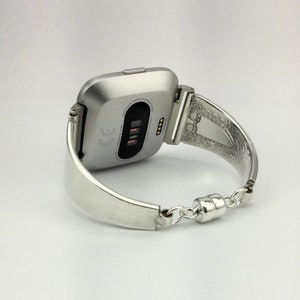 K&ouml;nnte beinhalten: Eine silberne Fitbit Smartwatch mit einem silbernen Metallarmband in Form einer Manschette. Das Zifferblatt der Uhr ist sichtbar und zeigt die Uhrzeit und das Datum an.