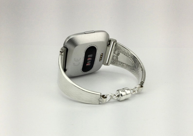K&ouml;nnte beinhalten: Eine silberne Fitbit Smartwatch mit einem silbernen Metallarmband in Form einer Manschette. Das Zifferblatt der Uhr ist sichtbar und zeigt die Uhrzeit und das Datum an.