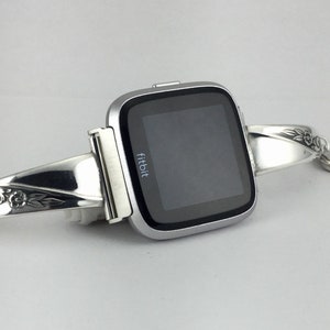 May include: A silver Fitbit smartwatch with a decorative silver bracelet. The bracelet has an engraved floral design.