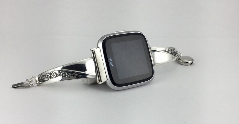 May include: A silver Fitbit smartwatch with a decorative silver bracelet. The bracelet has an engraved floral design.