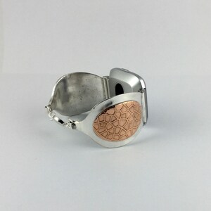 May include: A silver bracelet with a copper-coloured oval design. The bracelet is open and has a hinge on one side.