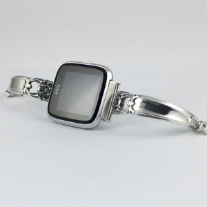 May include: A silver Fitbit smartwatch with a custom-made silver spoon band. The band is attached to the watch with a silver clasp.