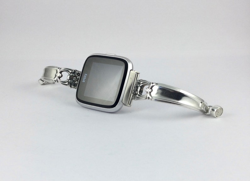 May include: A silver Fitbit smartwatch with a custom-made silver spoon band. The band is attached to the watch with a silver clasp.