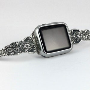 May include: A silver Apple Watch with a decorative, ornate silver band. The band is detailed with floral patterns.