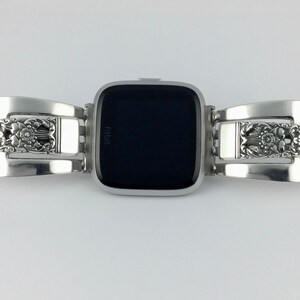 May include: Silver watch band made from repurposed silverware with an ornate floral design. The band is attached to a black Fitbit smartwatch.
