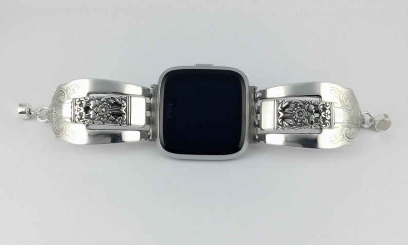 May include: Silver watch band made from repurposed silverware with an ornate floral design. The band is attached to a black Fitbit smartwatch.