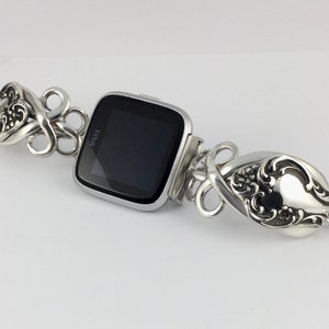 May include: Silver watch band made from repurposed silverware with a black Fitbit watch face. The band is made of two spoons with ornate designs.
