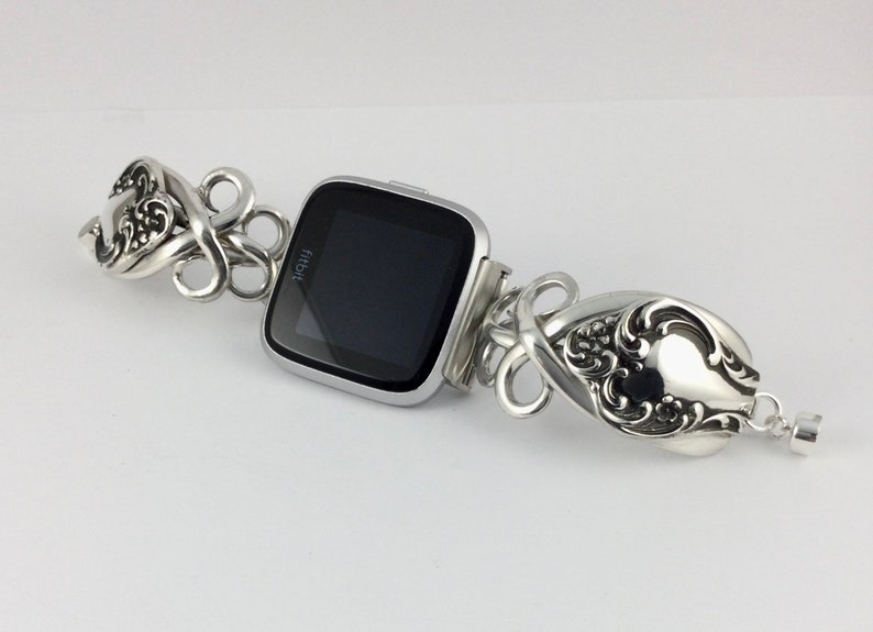 May include: Silver watch band made from repurposed silverware with a black Fitbit watch face. The band is made of two spoons with ornate designs.