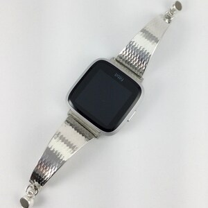 K&ouml;nnte beinhalten: Silberne Smartwatch mit einem strukturierten silbernen Armband. Das Zifferblatt der Uhr zeigt das Wort "Fitbit" an.