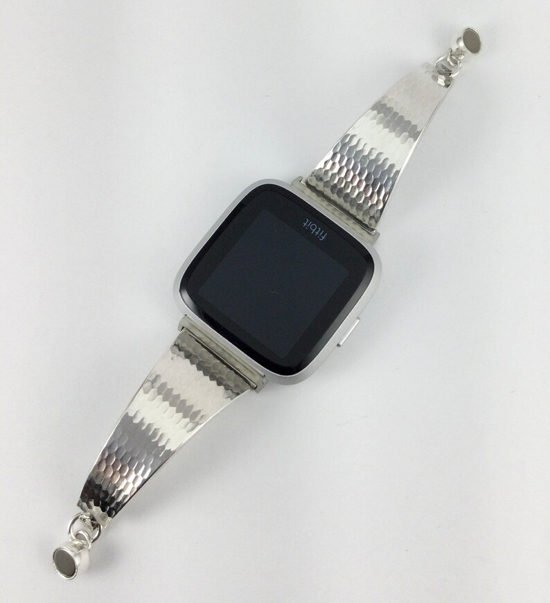 K&ouml;nnte beinhalten: Silberne Smartwatch mit einem strukturierten silbernen Armband. Das Zifferblatt der Uhr zeigt das Wort "Fitbit" an.