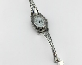 Könnte beinhalten: Eine silberfarbene Armreif-Uhr mit ovalem Zifferblatt, schwarzen arabischen Ziffern und dem Schriftzug Geneva Quartz. Das Armband ist mit detaillierten Schnörkelgravuren und einer Umrandung aus kleinen dunklen Steinen versehen.