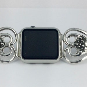 May include: Silver-toned watch band with a decorative floral design. The band is made of ornate metalwork, with a central section to hold the watch face. The design features floral and scroll motifs, creating an elegant and vintage aesthetic.