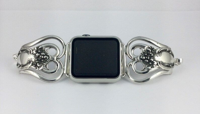 May include: Silver-toned watch band with a decorative floral design. The band is made of ornate metalwork, with a central section to hold the watch face. The design features floral and scroll motifs, creating an elegant and vintage aesthetic.