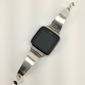 K&ouml;nnte beinhalten: Eine silberne Fitbit Smartwatch mit einem strukturierten Metallband. Das Zifferblatt der Uhr ist schwarz und zeigt das Wort "Fitbit" an.