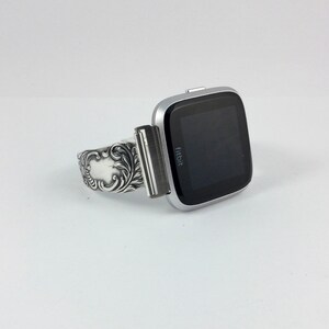 Puede incluir: Un reloj inteligente Fitbit plateado con una banda plateada decorativa. La esfera del reloj es negra y muestra la palabra "Fitbit".