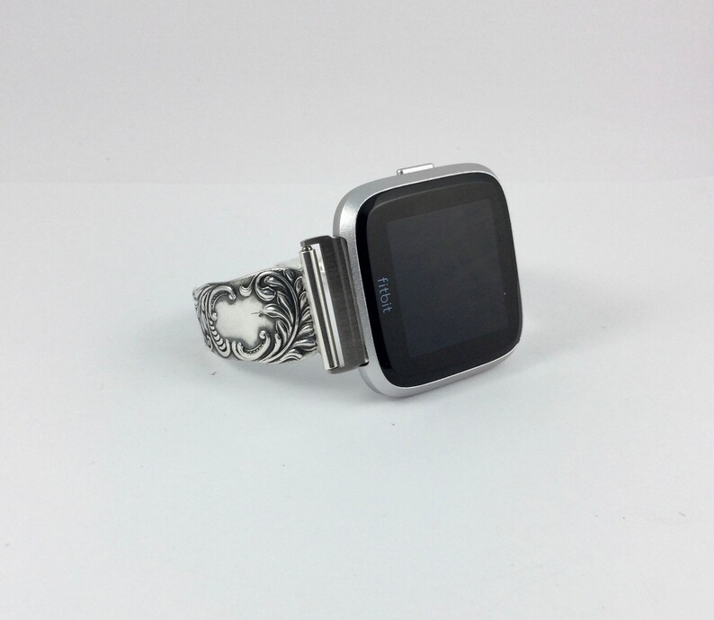 Puede incluir: Un reloj inteligente Fitbit plateado con una banda plateada decorativa. La esfera del reloj es negra y muestra la palabra "Fitbit".