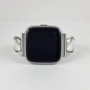 May include: A silver Fitbit smartwatch with a black screen. The watch band is made of two silver spoons.