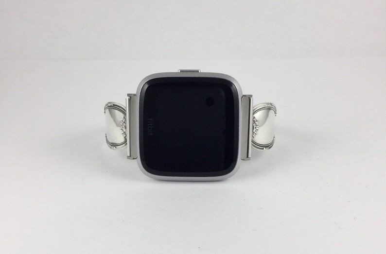 May include: A silver Fitbit smartwatch with a black screen. The watch band is made of two silver spoons.