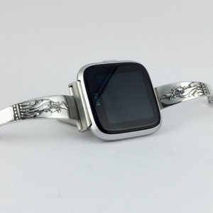 Op de afbeelding: Een zilveren Fitbit smartwatch met een zwart scherm is bevestigd aan een zilveren armband met een sierlijk ontwerp. De armband heeft een bloemmotief en is gemaakt van metaal.