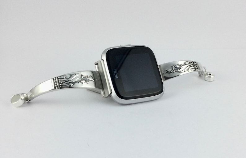 Op de afbeelding: Een zilveren Fitbit smartwatch met een zwart scherm is bevestigd aan een zilveren armband met een sierlijk ontwerp. De armband heeft een bloemmotief en is gemaakt van metaal.