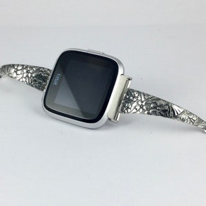 May include: A silver Fitbit smartwatch with a floral embossed band. The watch face is black and displays the word "fitbit".