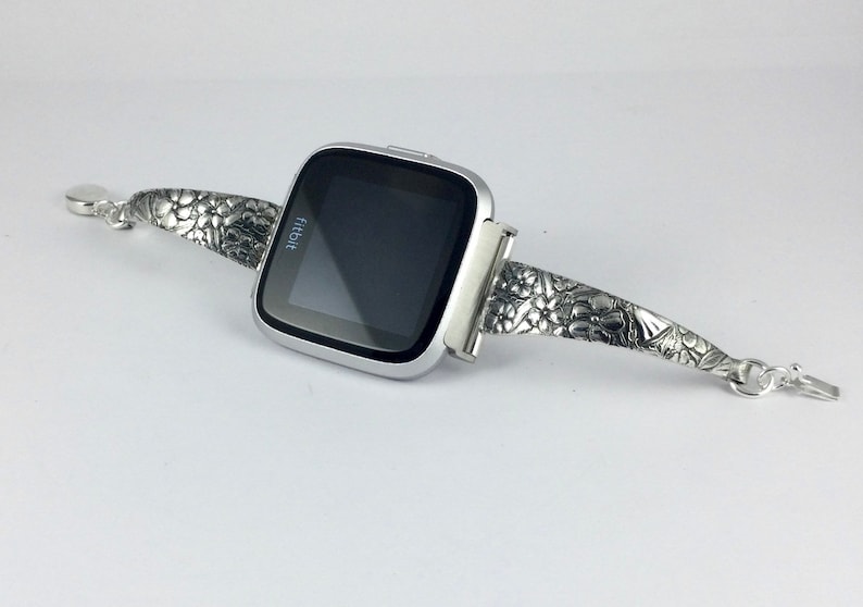 May include: A silver Fitbit smartwatch with a floral embossed band. The watch face is black and displays the word "fitbit".