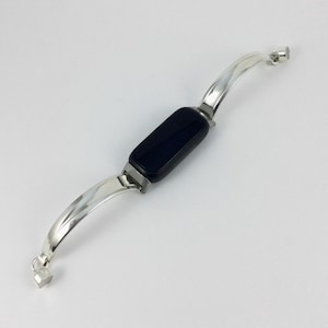 May include: A silver bracelet with a rectangular black center stone. The bracelet has a curved design with a clasp at each end. The metal has a polished finish, reflecting light.