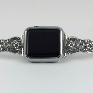 May include: Silver-toned Apple Watch band with an ornate floral design. The band features detailed carvings of flowers and leaves, adding a touch of elegance. The watch face is black.