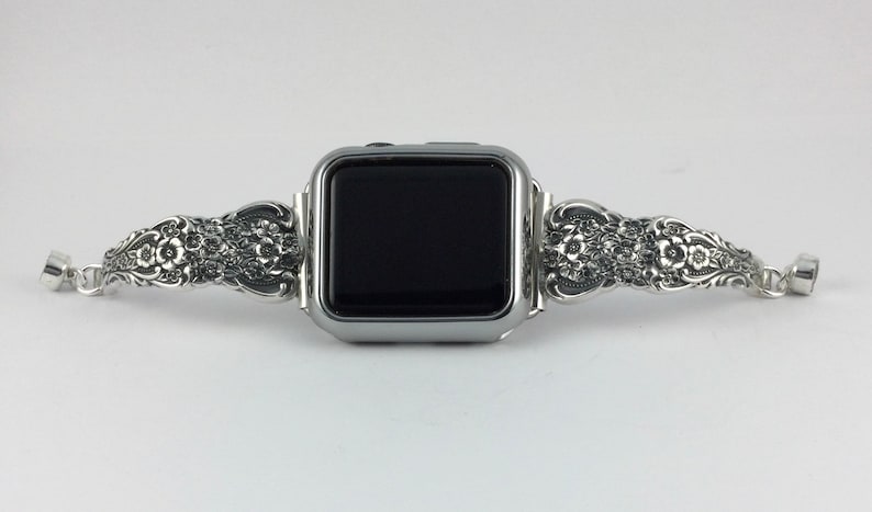 May include: Silver-toned Apple Watch band with an ornate floral design. The band features detailed carvings of flowers and leaves, adding a touch of elegance. The watch face is black.