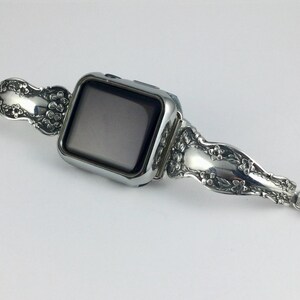 May include: A silver-toned smartwatch featuring a rectangular black screen and a decorative metal watch band. The band is crafted from repurposed silverware handles, showcasing an intricate embossed floral pattern and small circular magnetic clasps.