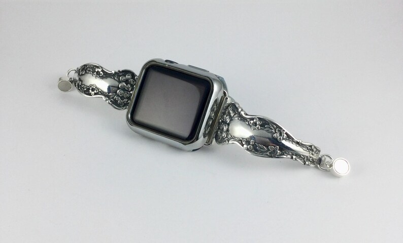 May include: A silver-toned smartwatch featuring a rectangular black screen and a decorative metal watch band. The band is crafted from repurposed silverware handles, showcasing an intricate embossed floral pattern and small circular magnetic clasps.