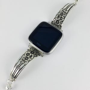 Pu&ograve; includere: Cinturino per orologio in argento con un design floreale. Il cinturino &egrave; attaccato a uno smartwatch Fitbit nero.