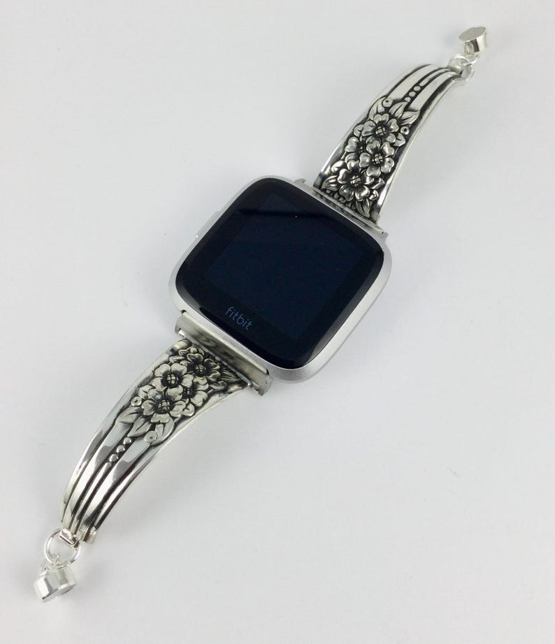 Pu&ograve; includere: Cinturino per orologio in argento con un design floreale. Il cinturino &egrave; attaccato a uno smartwatch Fitbit nero.