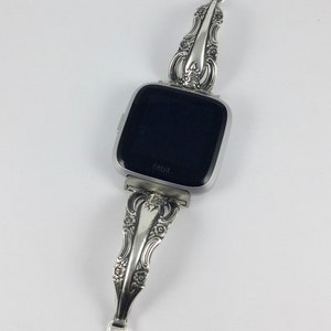 May include: A silver watch band made from a vintage silverware piece. The band is attached to a square-shaped smartwatch with a black screen that says "fitbit".