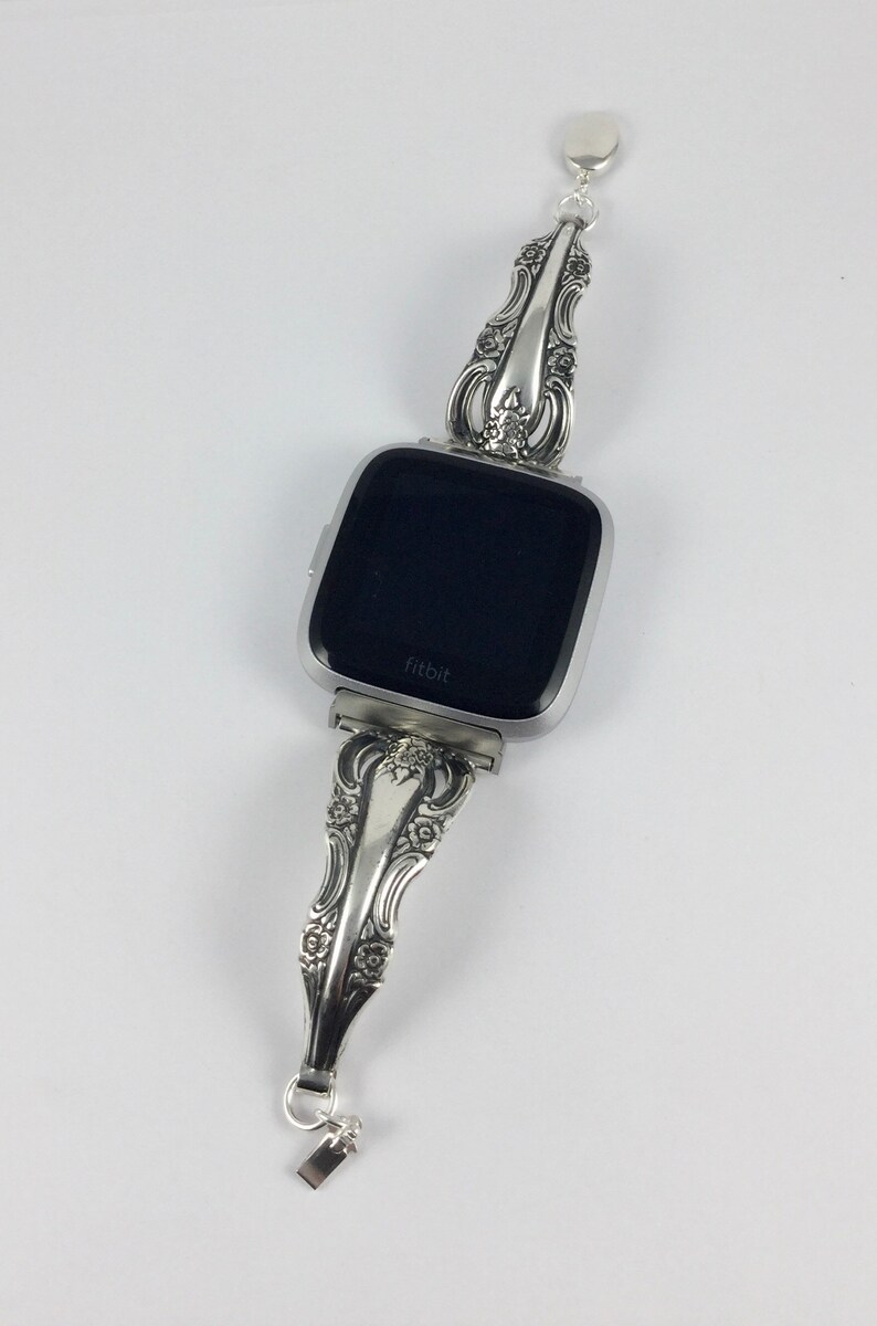 May include: A silver watch band made from a vintage silverware piece. The band is attached to a square-shaped smartwatch with a black screen that says "fitbit".