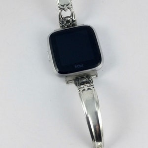 May include: Silver watch band made from a repurposed spoon with a black Fitbit watch face. The band has a floral design and a heart-shaped charm.