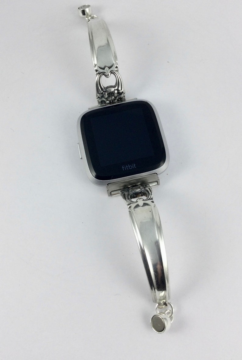 May include: Silver watch band made from a repurposed spoon with a black Fitbit watch face. The band has a floral design and a heart-shaped charm.