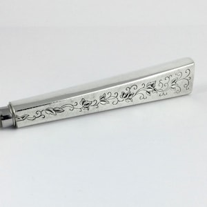 May include: A silver stylus with a floral design engraved on the body. The stylus has a rubber tip.