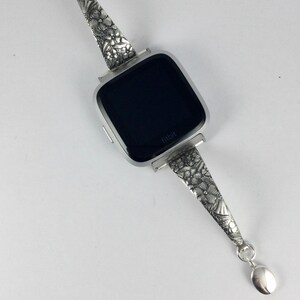 May include: A silver watch band with a floral design. The band is attached to a black Fitbit watch face.