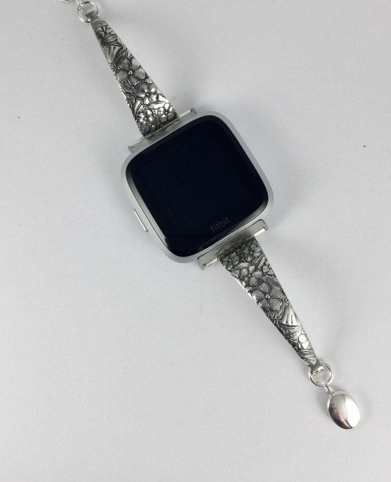 May include: A silver watch band with a floral design. The band is attached to a black Fitbit watch face.