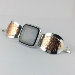 May include: A silver and rose gold watch band with a textured design. The watch face is black and displays the word "Fitbit".