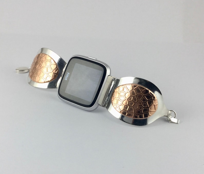 May include: A silver and rose gold watch band with a textured design. The watch face is black and displays the word "Fitbit".