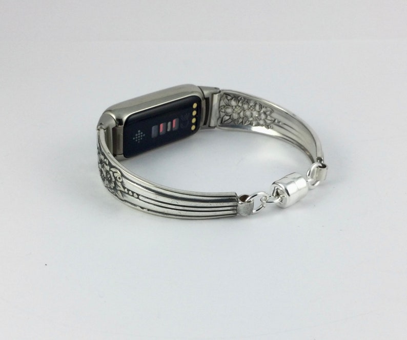 Może przedstawiać: Srebrny pasek do zegarka Fitbit wykonany z przetworzonych sztućc&oacute;w. Pasek ma skomplikowane wzory kwiatowe i liniowe, z magnetycznym zapięciem. Tarcza zegarka wyświetla czarny ekran z kolorowymi wskaźnikami.