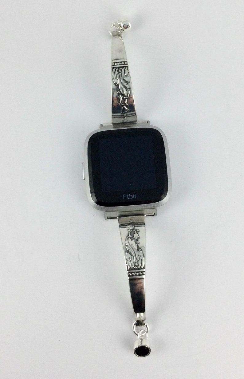 Op de afbeelding: Zilveren horlogeband met een sierlijk ontwerp, met een zwarte Fitbit smartwatch met het woord "fitbit" op het display.